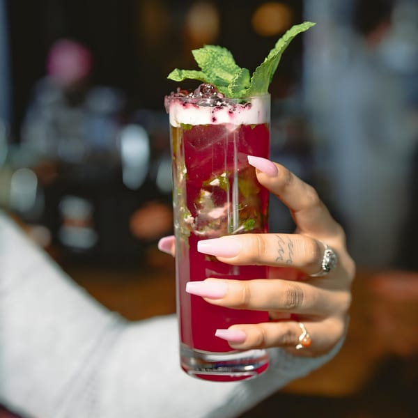 A hand with pink nails holds a tall glass of red cocktail garnished with mint and berries.