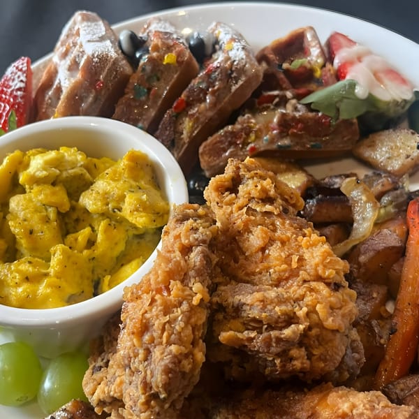 A soul food platter with fried chicken, scrambled eggs, waffles, fruit, and roasted vegetables.