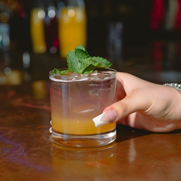 A hand with a diamond bracelet holding a cocktail garnished with mint on a wooden bar.