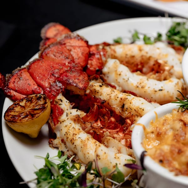 Grilled lobster tails with a charred lemon, greens, and a side of macaroni and cheese.
