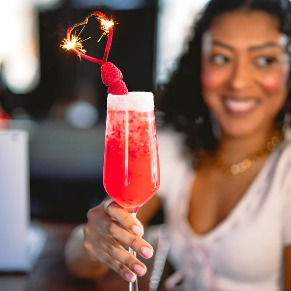 A woman holding a vibrant red cocktail garnished with raspberries and a heart-shaped sparkler.