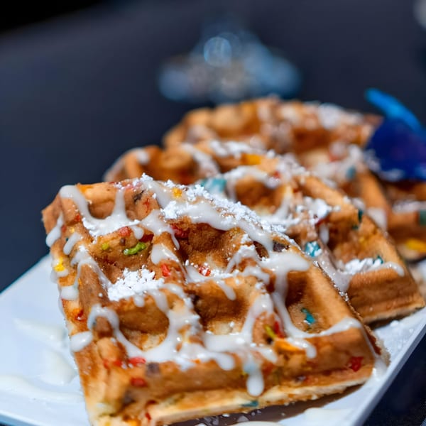 Close-up of golden-brown waffles with colorful sprinkles, drizzled with white icing and powdered sugar.