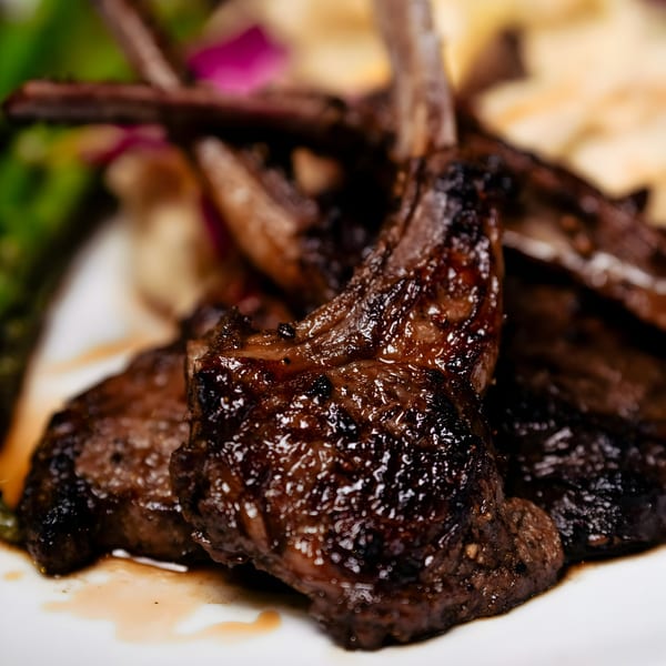 Close-up of glistening, grilled lamb chops with bones, served on a white plate at Noir Kitchen & Cocktails.