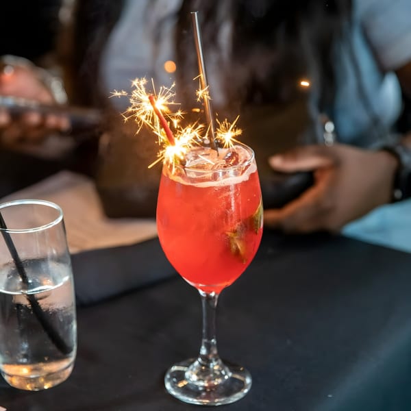 A vibrant red cocktail with sparklers and a metal straw, next to a glass of water on a dark table.