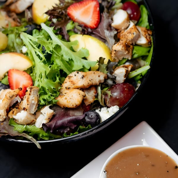 A vibrant salad with grilled chicken, mixed greens, strawberries, apples, grapes, blueberries, and feta cheese, with dressing on the side.