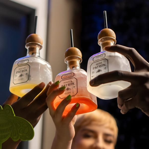 Three hands holding up Patron tequila bottles filled with colorful cocktails, ready for a toast.