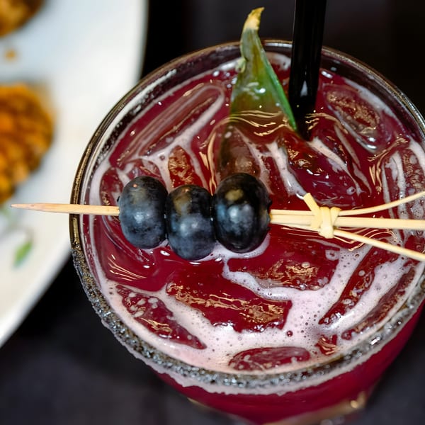 Close-up of a vibrant red cocktail with ice, garnished with three blueberries and a green leaf.