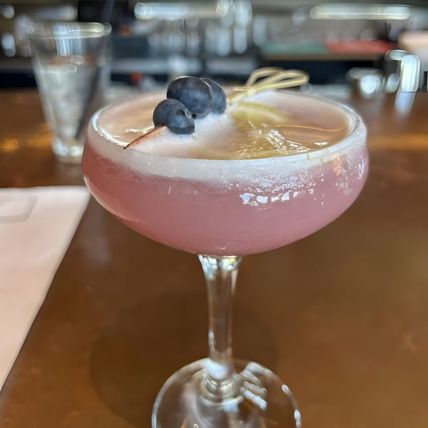 A pink cocktail in a coupe glass, garnished with blueberries and a lemon peel, on a bar.